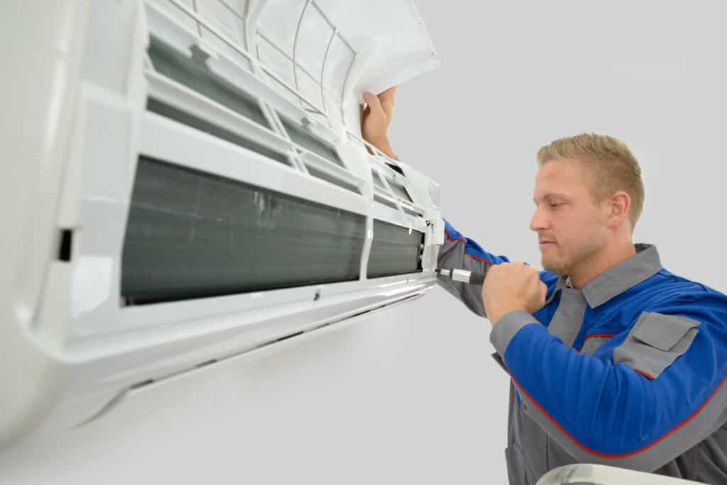 Technician in a blue and gray uniform inspects and repairs a wall-mounted air conditioning unit, lifting the front panel to access the interior components.