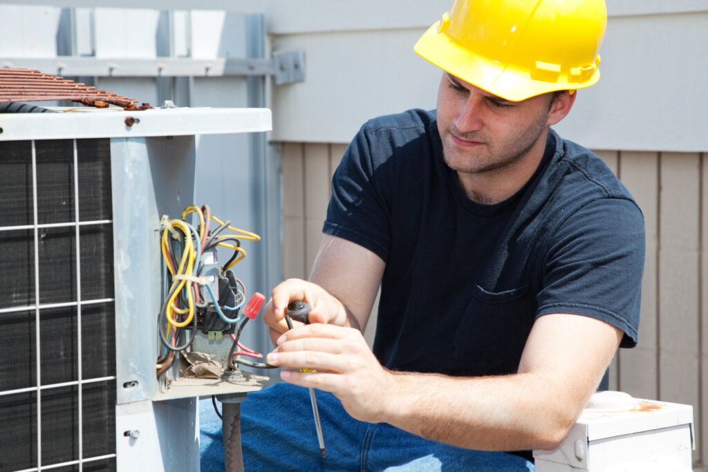 A worker in a yellow hard hat uses tools to repair an air conditioning unit.