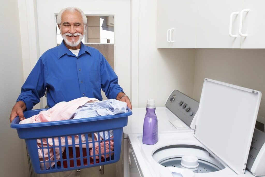 Man smiling, holding a laundry basket with clothes. A washing machine with an open lid and a bottle of detergent are nearby.