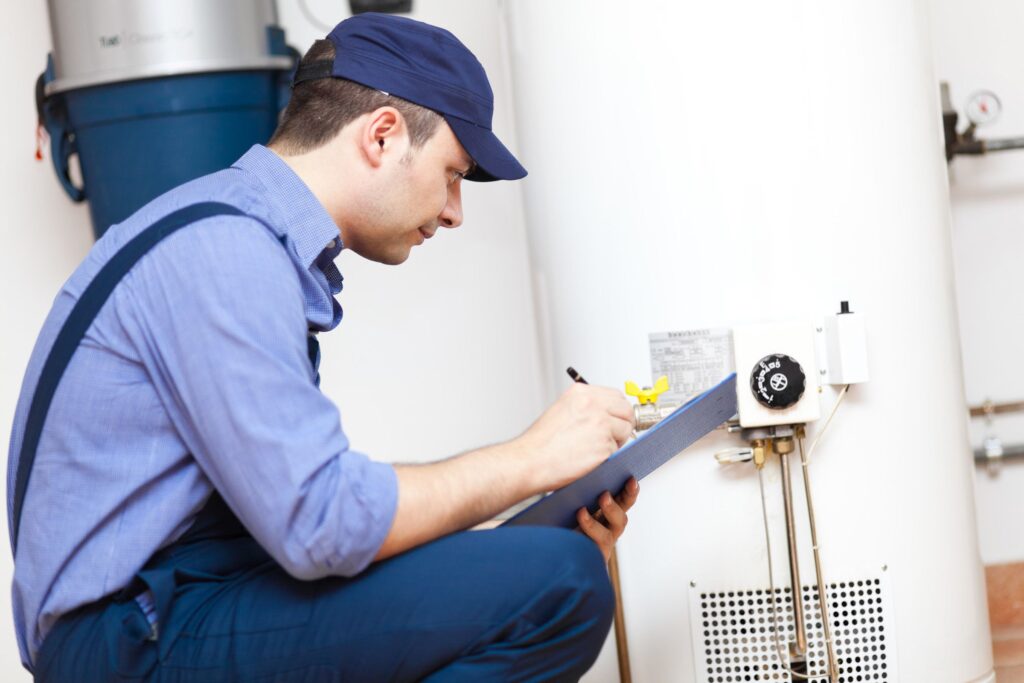 A technician in blue uniform checks and writes on a clipboard while inspecting a white water heater in a utility room.