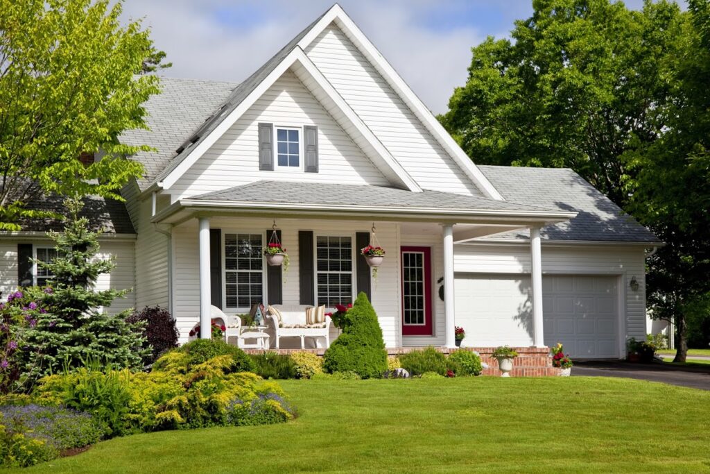 A white suburban house with a gabled roof, front porch, and attached garage. The lawn is well-maintained with shrubs and hanging flower baskets.