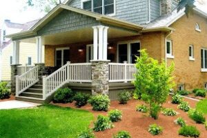Front view of a house with gray and brick exterior, featuring a porch with stone pillars and railing. The landscaped yard has green shrubs and mulch.