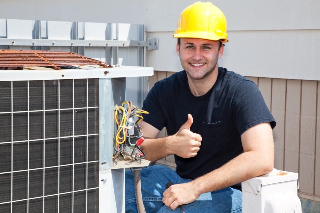 A worker in a yellow hard hat sits next to an HVAC unit, smiling and giving a thumbs-up.