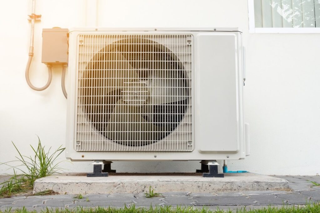 Close-up of an outdoor air conditioning unit positioned on a concrete slab next to a building wall, with grass and a small pathway visible in the foreground.