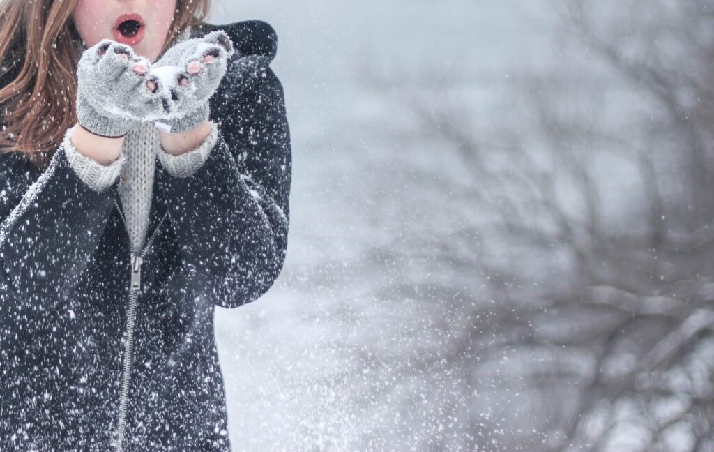A HELP team member in a dark coat playfully blows snow from their hands outside, with snowy trees softly blurred behind them.