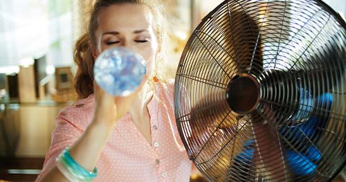 Image: a young woman drinks from a water bottle next to a tabletop fan. For the best Centerville air conditioning services, call HELP.