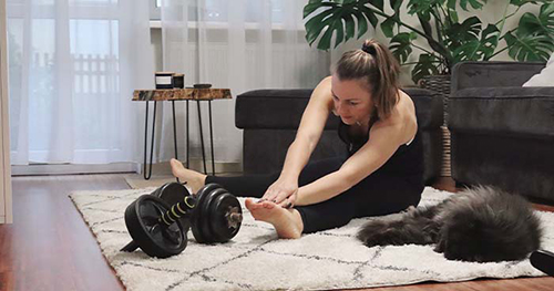 Image: a woman stretching with her cat before starting a workout in her home gym.