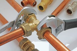 Two wrenches adjust a brass pipe fitting connecting copper pipes, surrounded by various plumbing connectors on a white background.