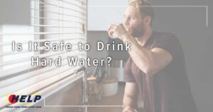 A man stands by a window sipping a glass of water, contemplating the question, "Is It Safe to Drink Hard Water?" The image features a plumbing service logo.