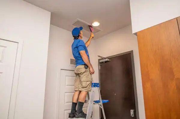 A person in a blue shirt and shorts stands on a ladder, cleaning a ceiling air vent with a tool inside a room.