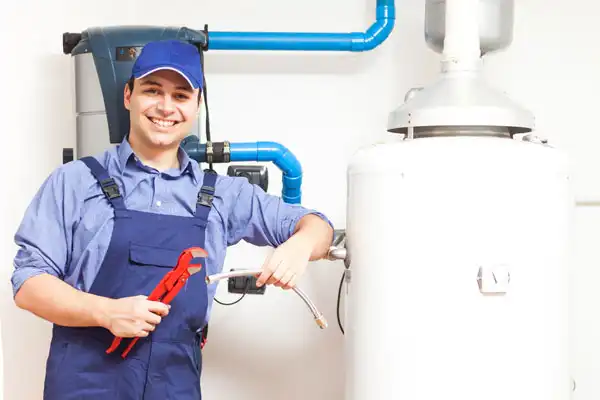 A plumber in blue overalls and cap holding a red pipe wrench and a flexible pipe stands next to a water heater, smiling at the camera.