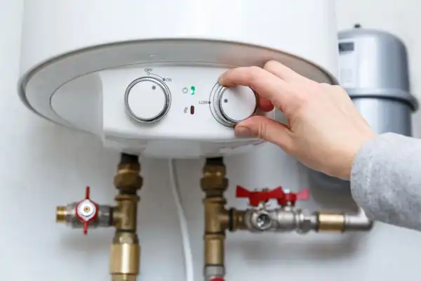 Hand adjusting the temperature dial on a water heater with visible pipes and valves below.