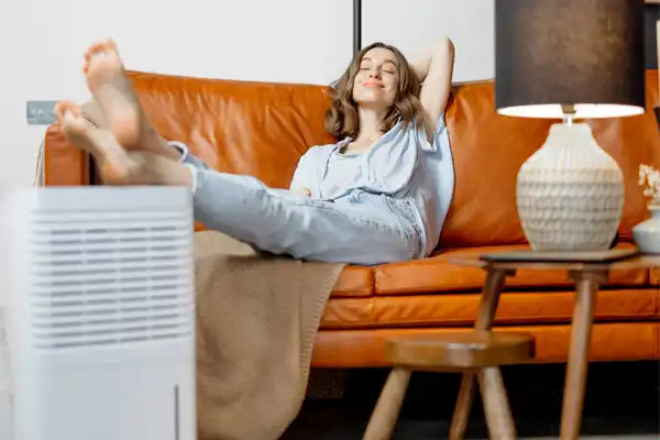 A person relaxes on a brown leather sofa with feet up, eyes closed, near a small white appliance and a lamp on a wooden side table.