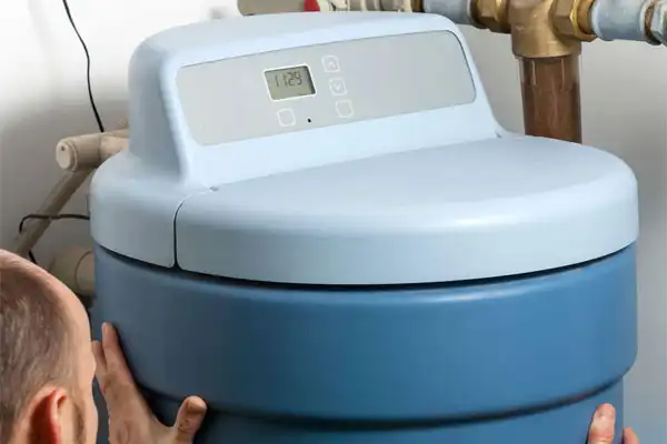 A person adjusts a blue water softener unit with control buttons and digital display on top. Copper pipes are visible in the background.