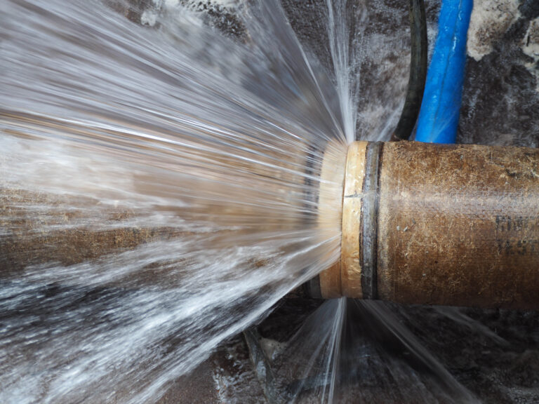 Water is forcefully spraying from a cracked section of a brown pipe, causing a leak. Blue tubing is visible in the background.