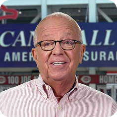 A smiling older man in glasses and a striped shirt stands in front of a blurred background with signage.