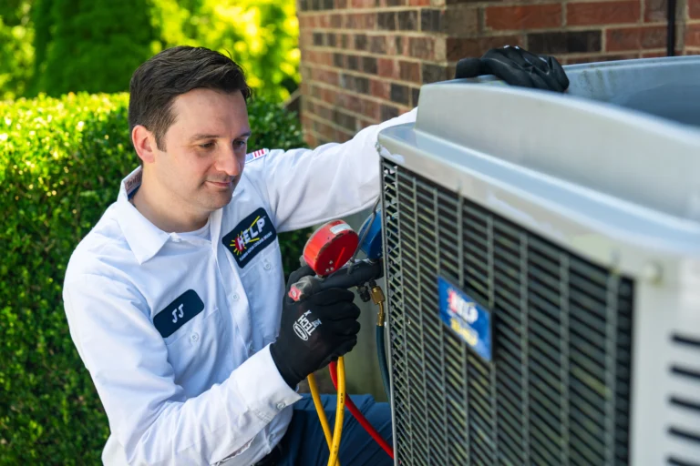 A HELP team technician in a white uniform and black gloves services an outdoor AC unit beside a brick wall.