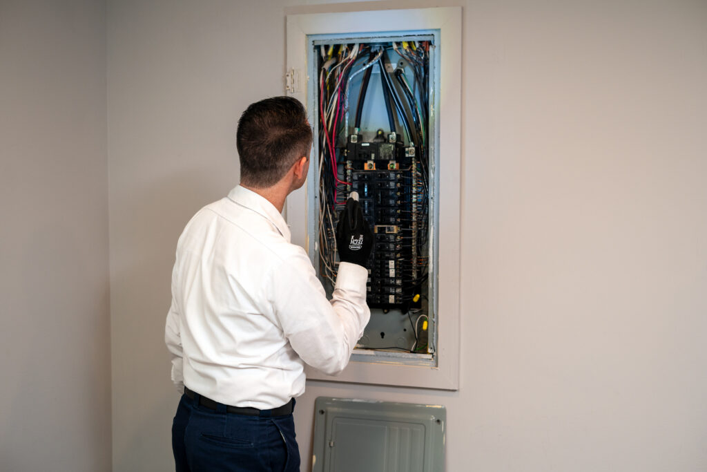 A HELP team member in a white shirt carefully examines an open electrical panel with multiple wires, wearing a protective glove.