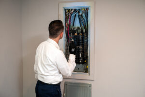 A HELP team member in a white shirt carefully examines an open electrical panel with multiple wires, wearing a protective glove.