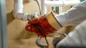 A HELP Plumbing technician in red gloves uses a plumbing snake to clear a clog in white PVC pipes beneath a customer’s sink.