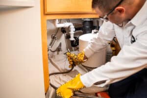 A HELP Plumbing, Heating, Cooling and Drains technician, wearing protective gloves, uses a plumbing snake beneath a kitchen sink to remove a stubborn clog. The cabinet contains various pipes, cleaning supplies, and helpful information about water heater replacement services available in Cincinnati.